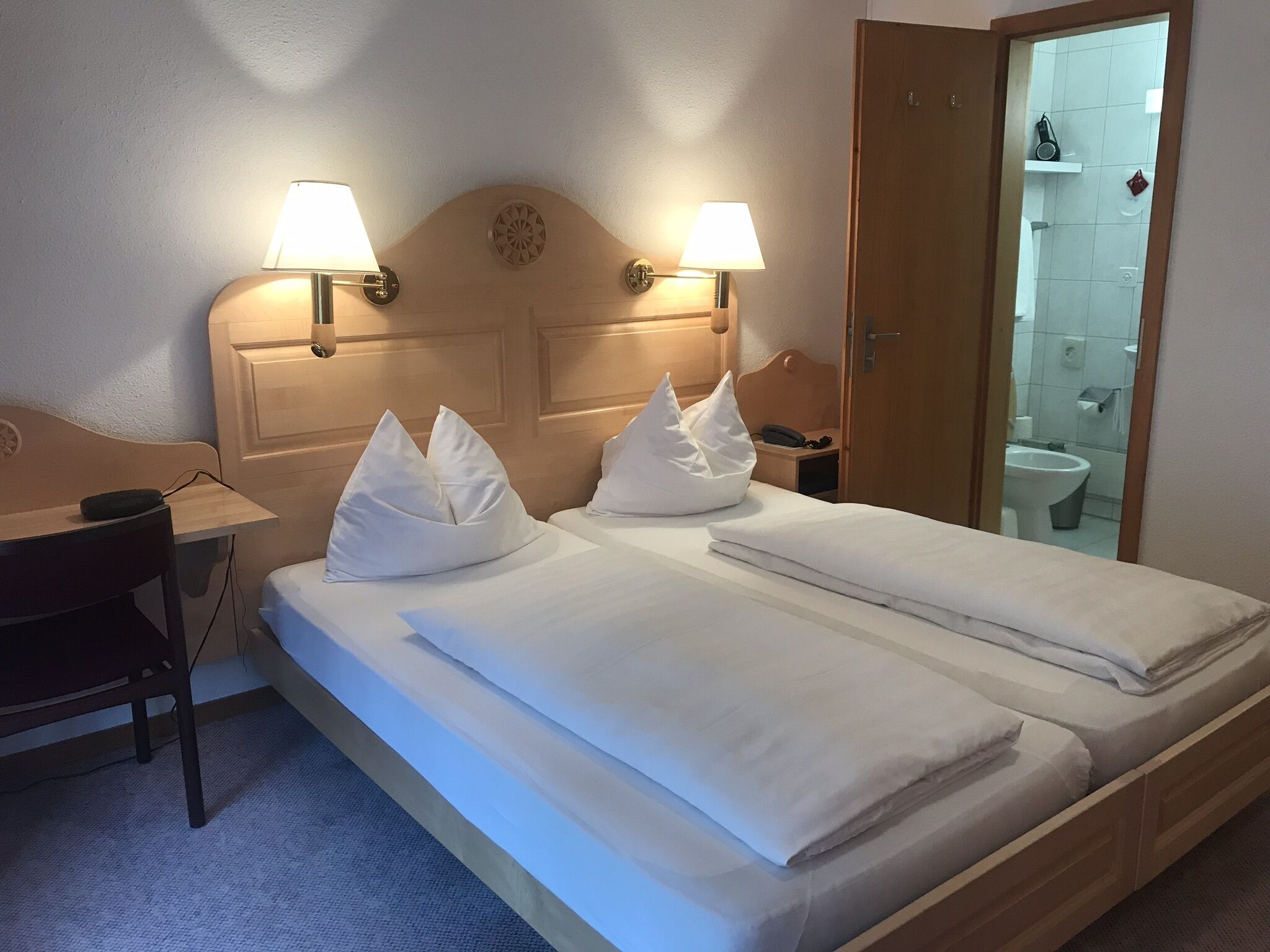 Ein Hotelzimmer mit Doppelbett, zwei Nachttischlampen, einem Holz-Kopfteil, einem Schreibtisch mit Stuhl und einem durchgehenden Türspalt sichtbaren Badezimmer.