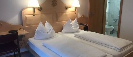 A hotel room with a double bed, two bedside lamps, a wooden headboard, a desk with a chair, and a bathroom visible through an open door.