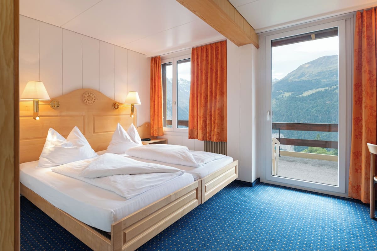 Ein Hotelzimmer mit einem Bett, zwei Nachttischlampen, einem Fenster mit Vorhängen und Blick auf Berge.