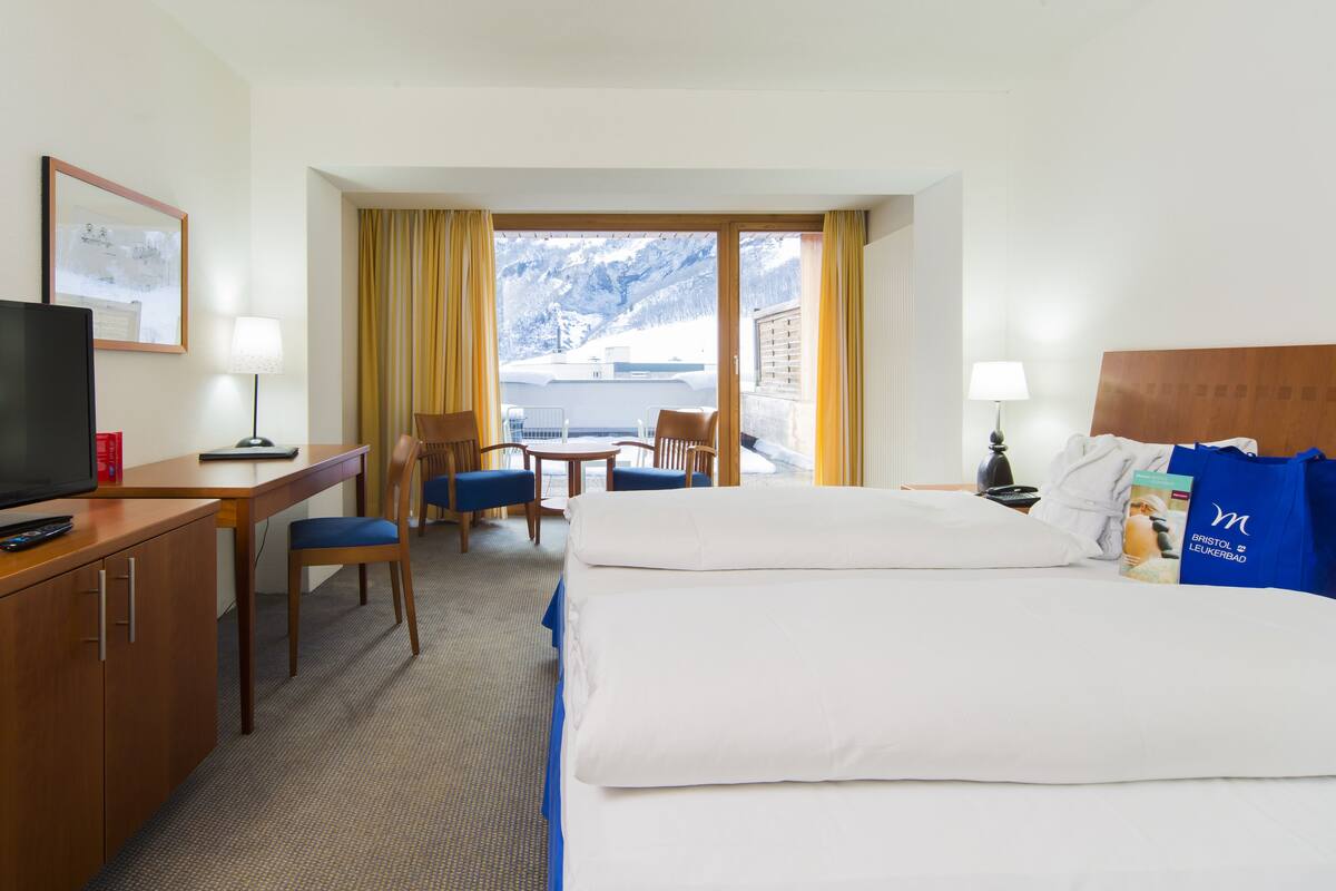 A hotel room with a bed, a desk with a television, a chair, a table, and a view of mountains through the window.