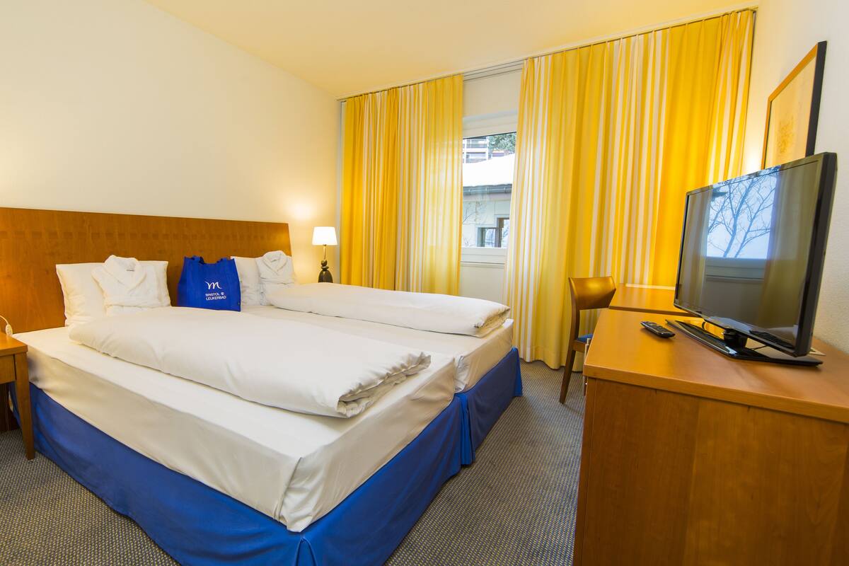 A hotel room with a large bed, two bedside tables, a TV, and a window with yellow curtains.
