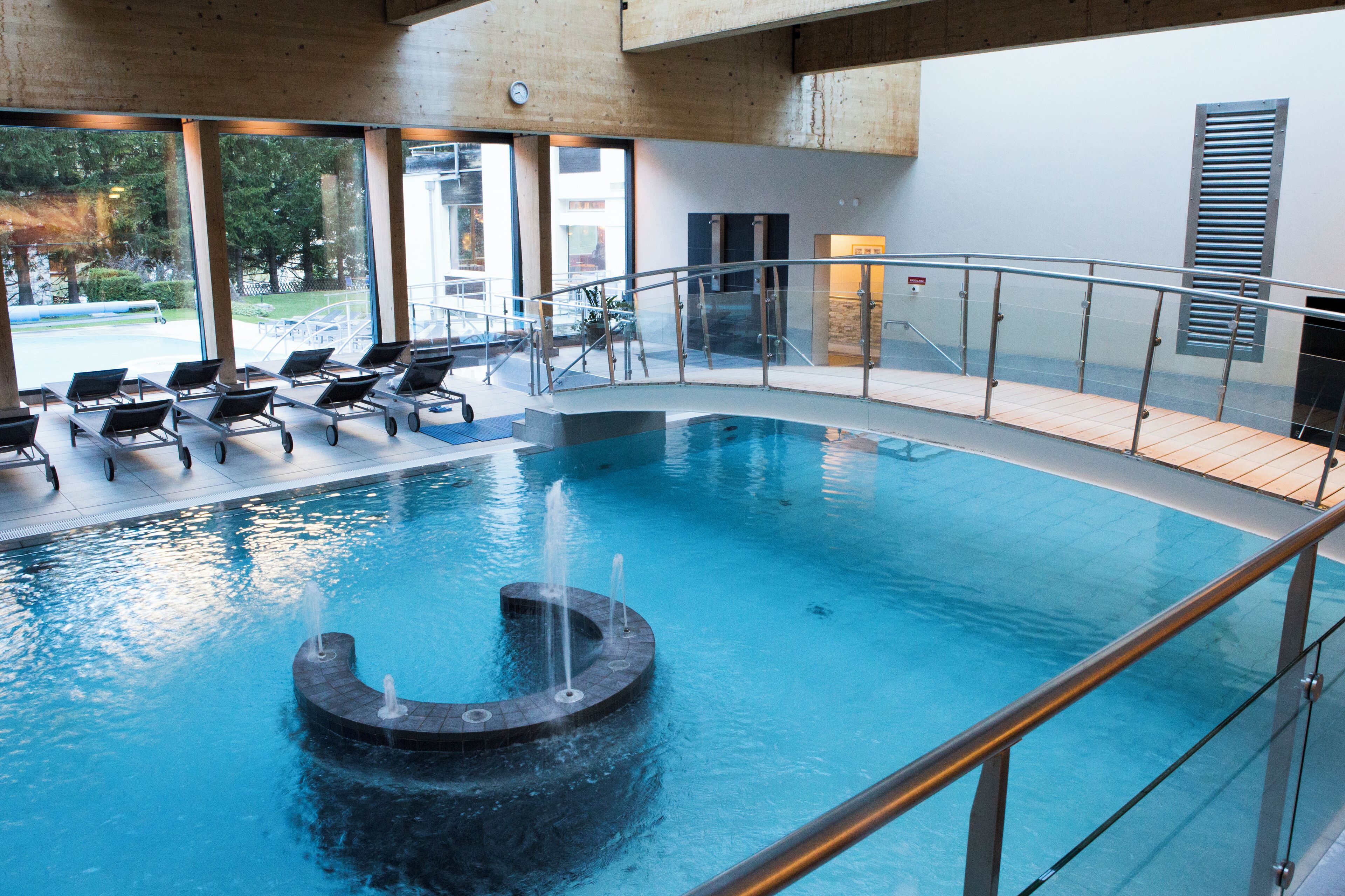 Ein moderner Innenpool mit einem runden Wasserspiel, Liegestühlen und einer Glasbrüstung.