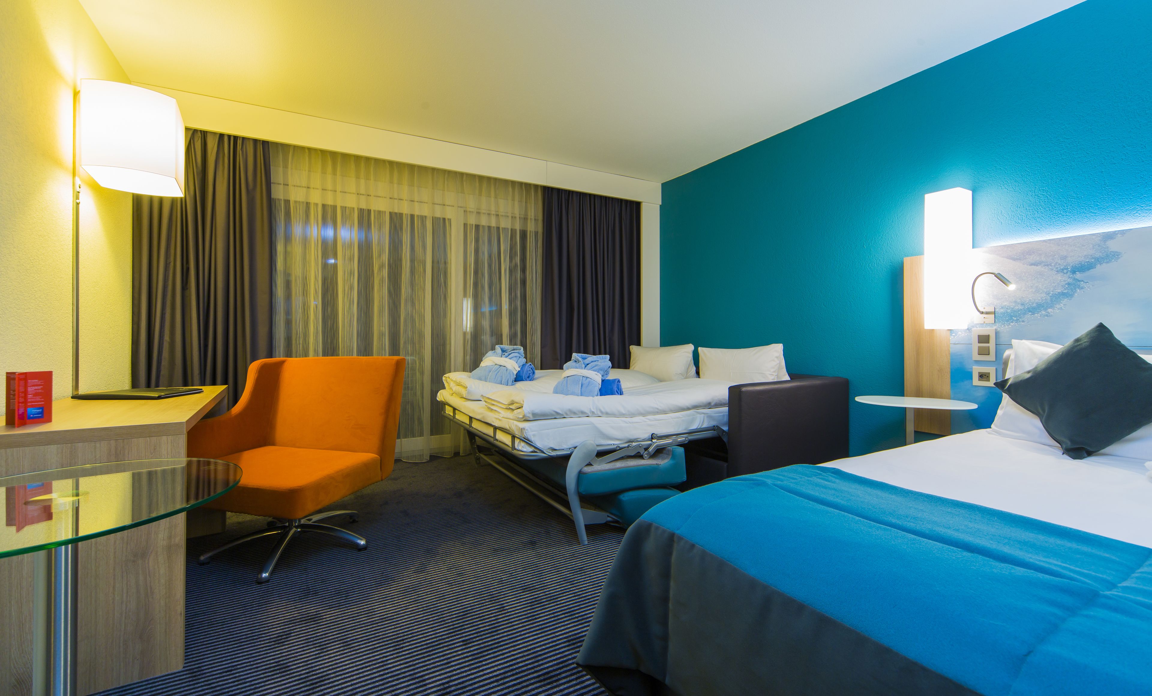 A hotel room with a bed, a desk, a chair, a bedside table, and a window with curtains.