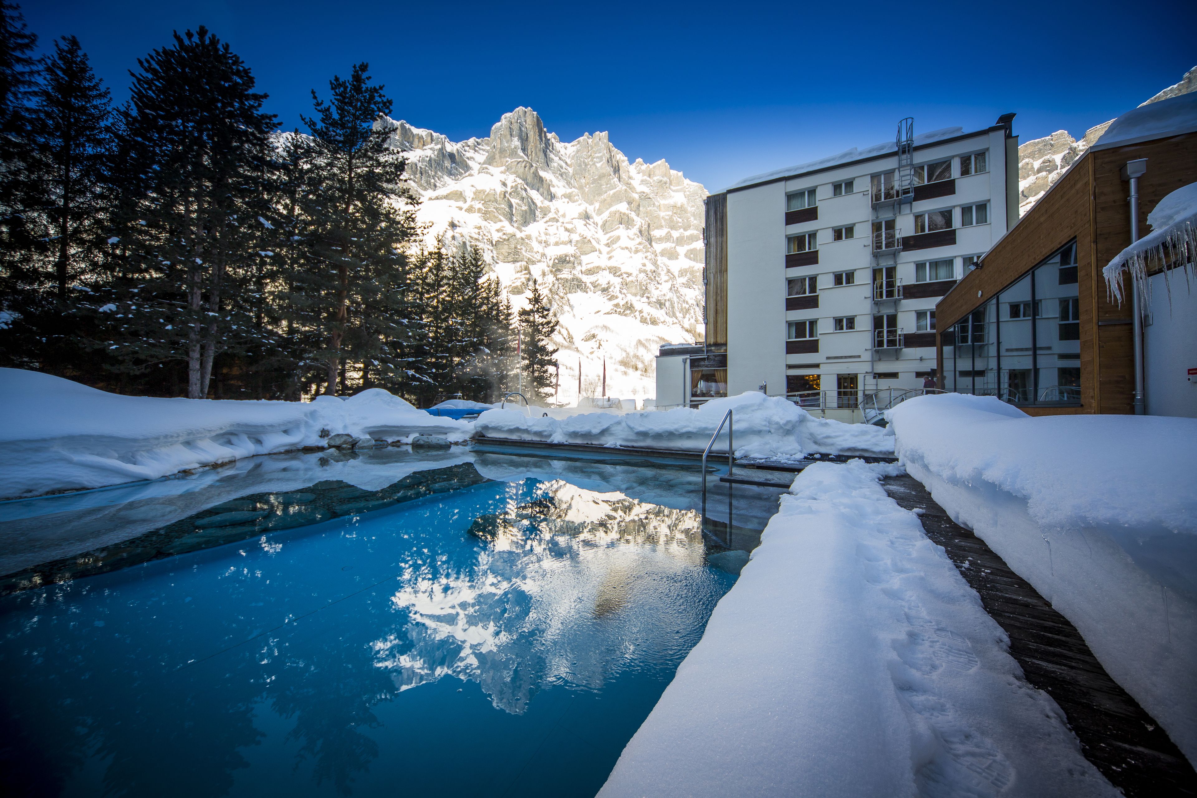 A pool surrounded by snow, with a mountain in the background and a building with balconies.