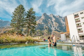 A swimming pool with a mountain backdrop, a wooden deck, and a modern building with balconies. - Le Bristol Leukerbad (Leukerbad)