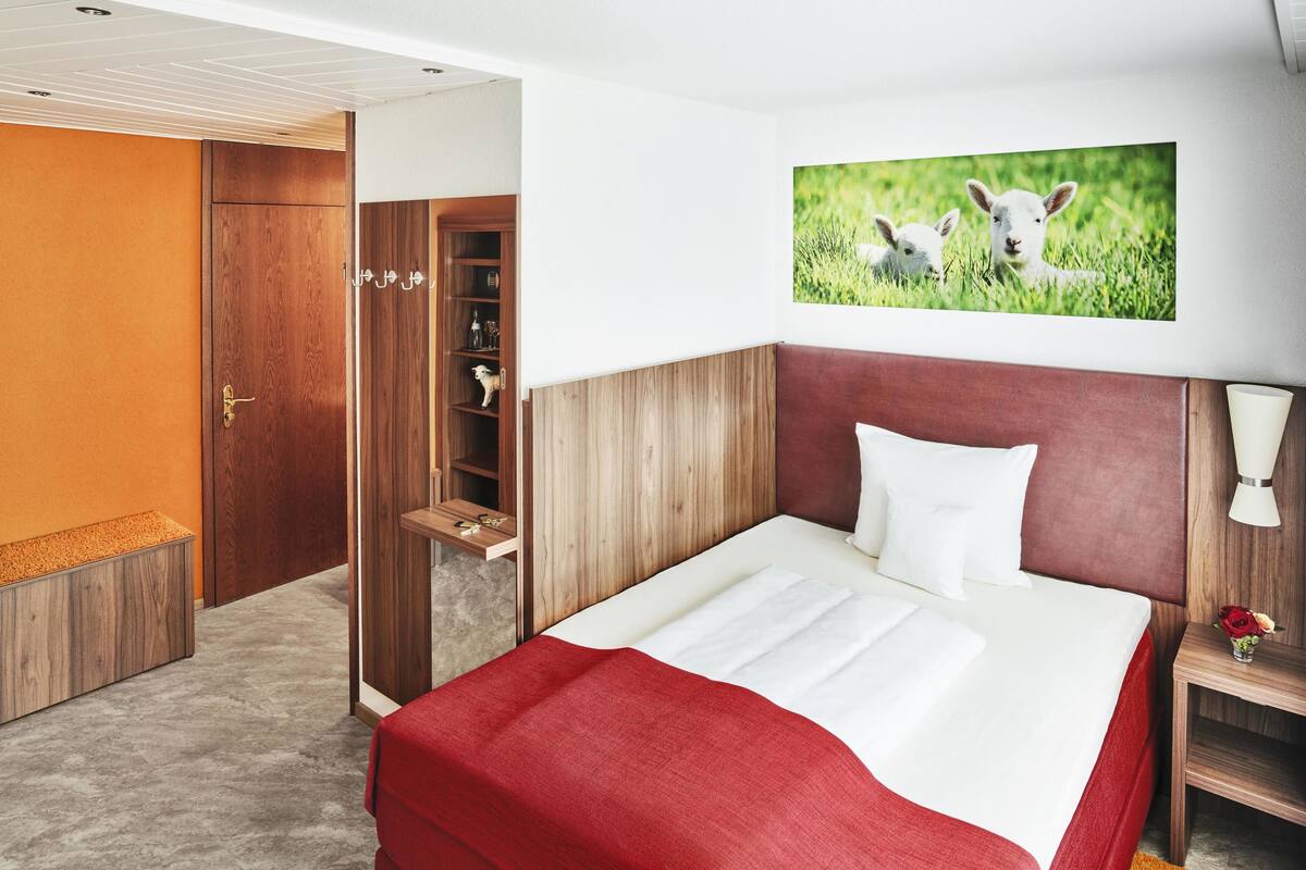 Ett hotellrum med en säng, en garderob i trä, en hylla med dekorativa föremål och en tavla med motiv av lamm som hänger på väggen.