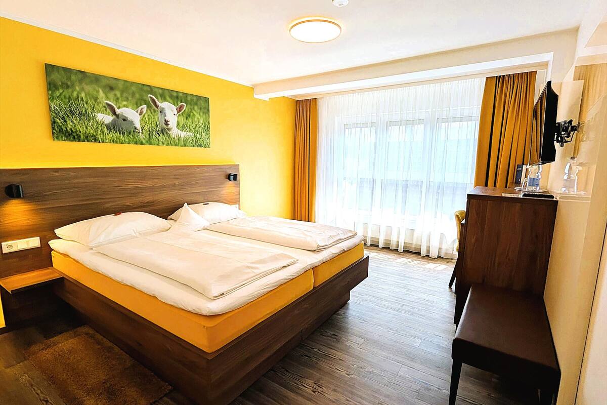 Ett hotellrum med en stor säng, ett skrivbord, en tv och en tavla med får på väggen.