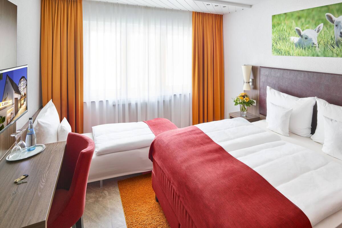 Ett hotellrum med två sängar, ett skrivbord, en stol, en tv och en tavla som föreställer lamm.