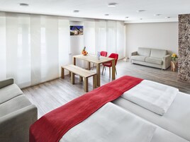 Una habitación de hotel moderna con cama, una manta roja, una zona de comedor con mesa y sillas, y un sofá.