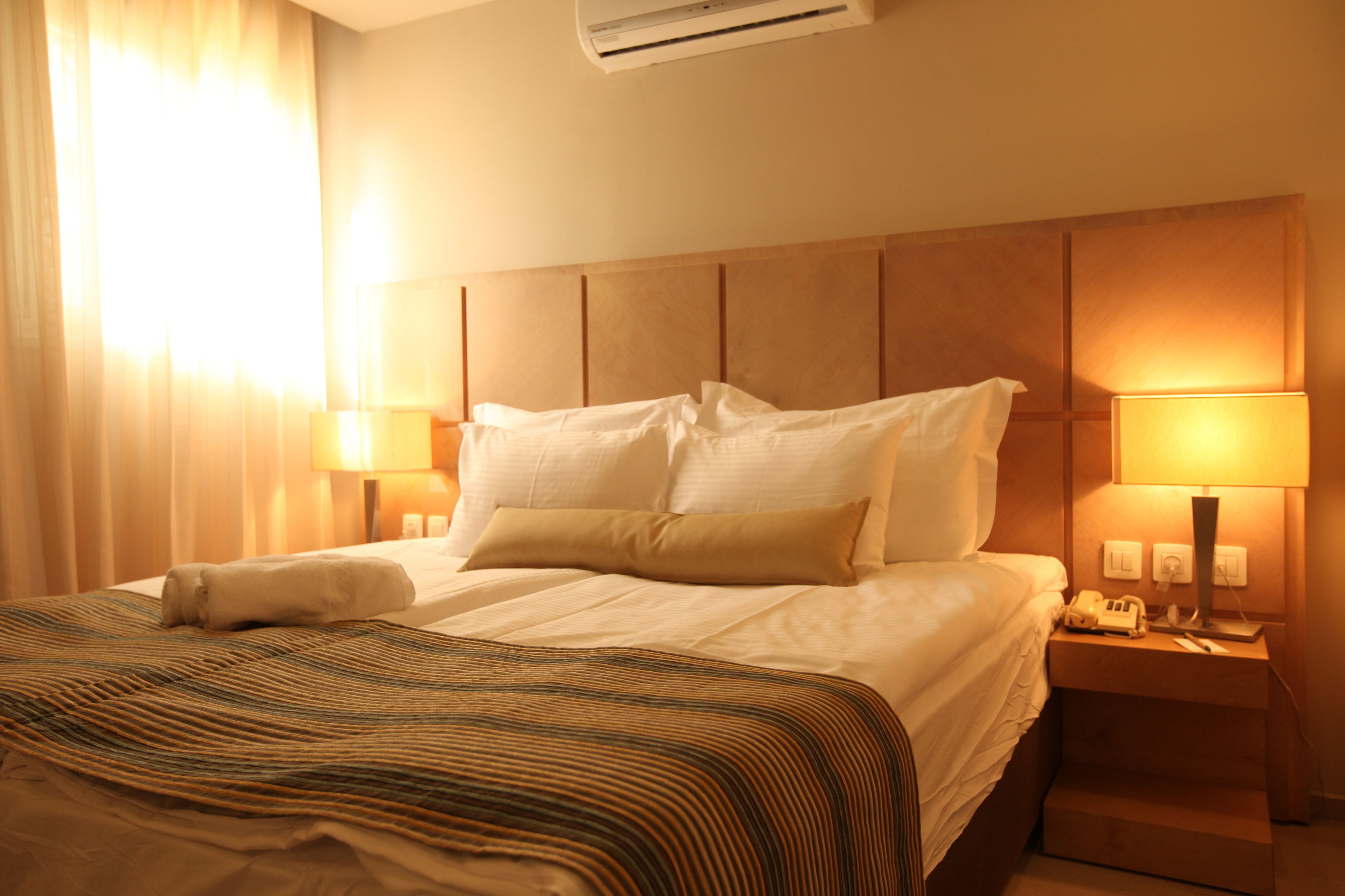 A hotel room with a large bed, two bedside lamps, a nightstand with a phone, and an air conditioning unit.