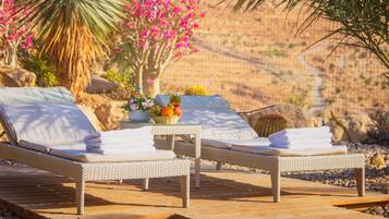 A wooden deck with two lounge chairs, a table with a fruit bowl, and a vase of flowers.