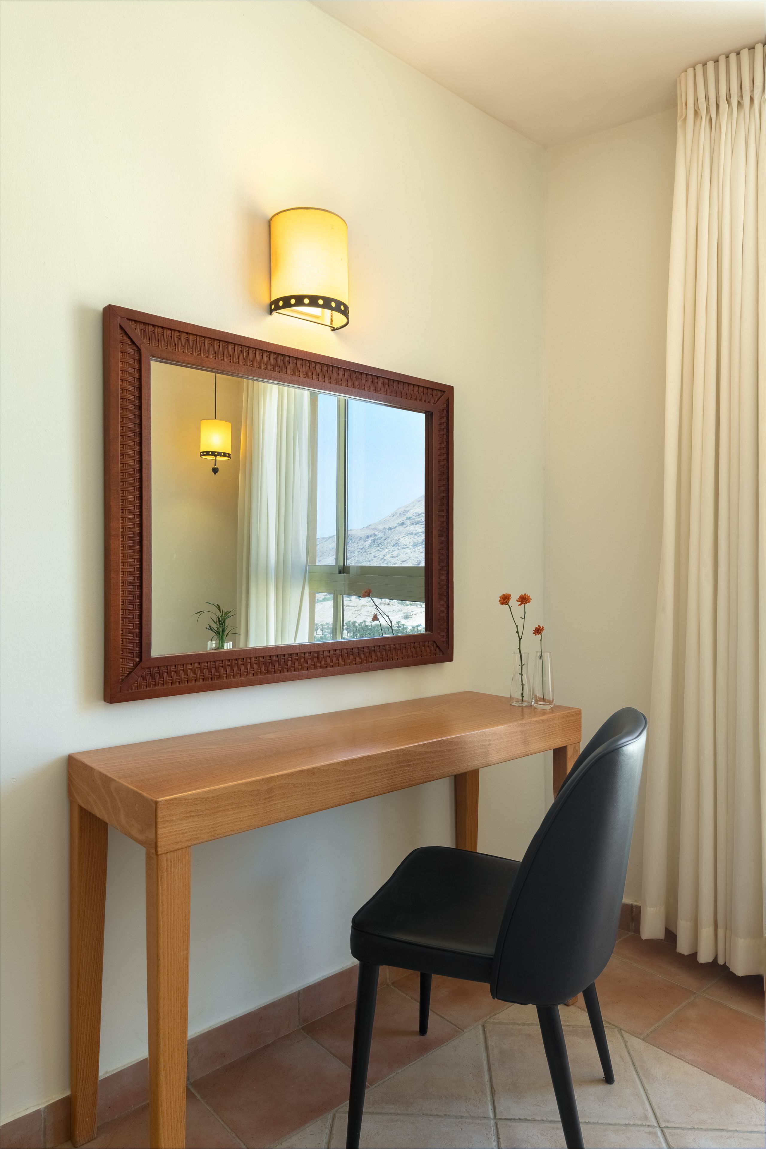 A wooden desk with a black chair, a mirror with a wooden frame, and a wall-mounted lamp.