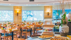 Un grand espace dédié à la salle à manger, avec de nombreuses tables et chaises, un buffet proposant une variété de plats et de grandes fenêtres laissant entrer la lumière naturelle.