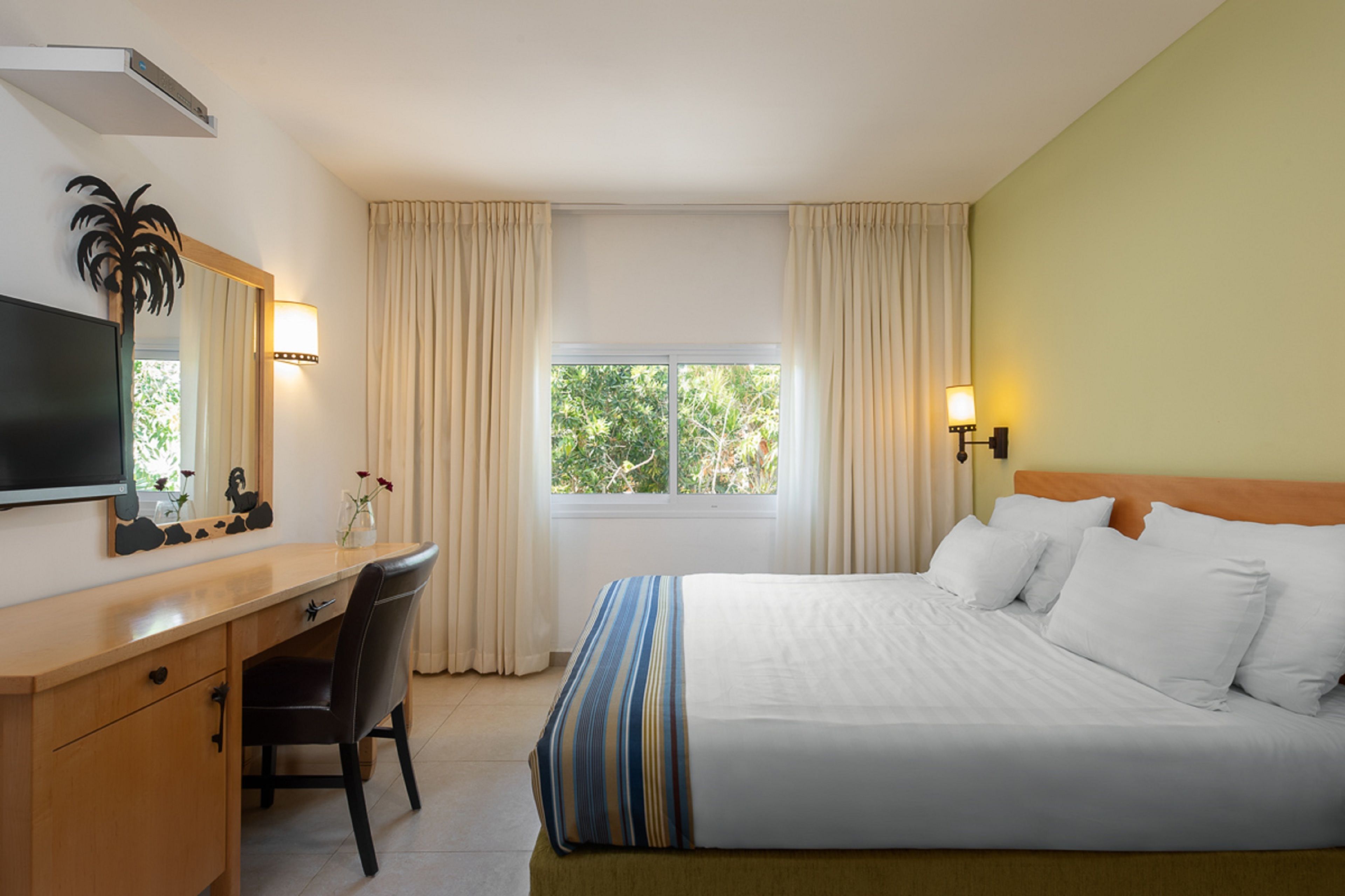 Una habitación de hotel con cama, escritorio, silla, televisor, ventana con cortinas y lámpara fijada a la pared.