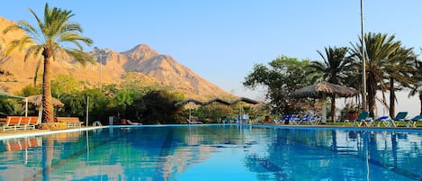 A swimming pool with lounge chairs and palm trees, set against a mountainous backdrop.
