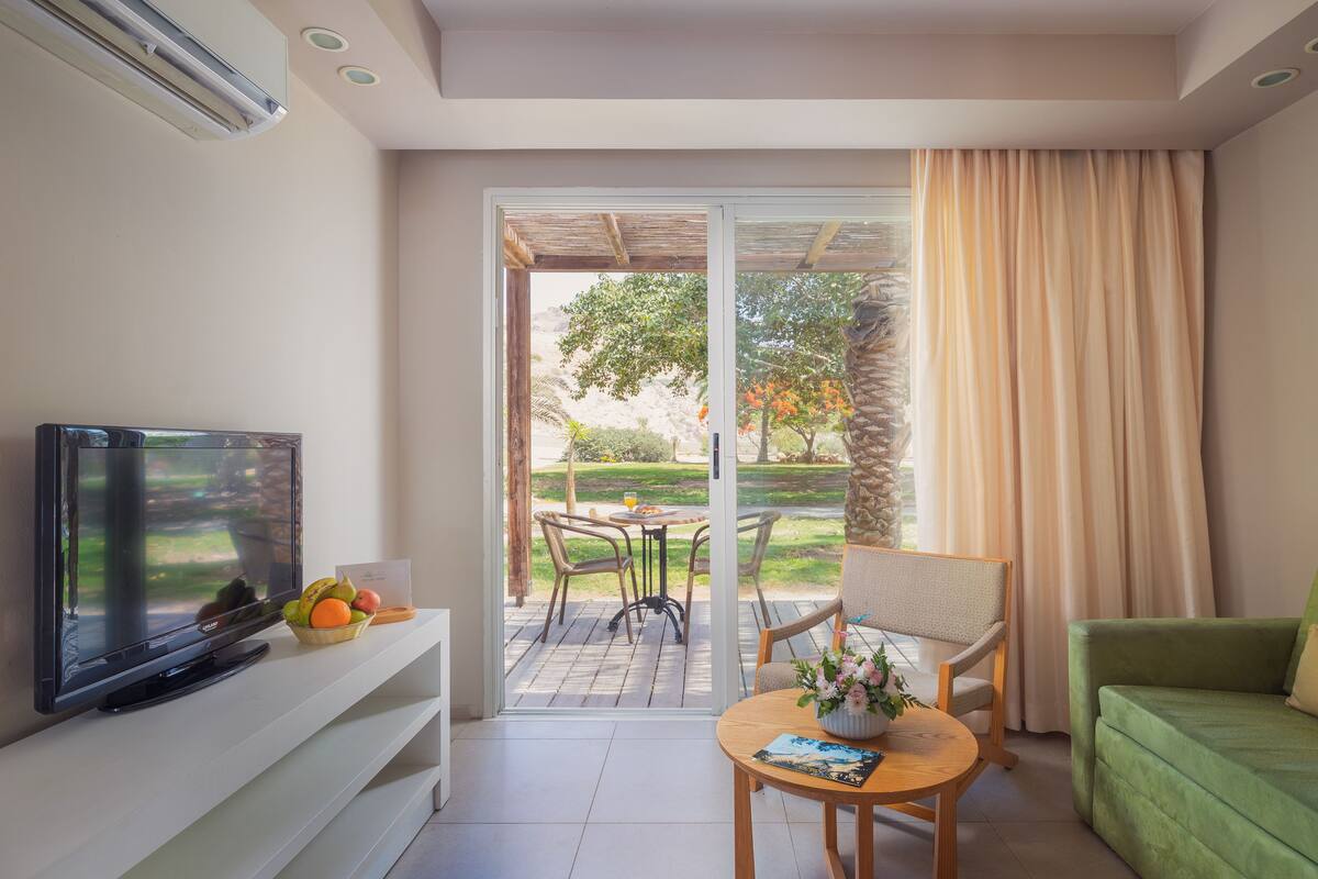 Un salon moderne avec un téléviseur à écran plat, une table basse en bois sur laquelle sont posés un livre et des fleurs, un canapé vert et une porte-fenêtre coulissante donnant sur une terrasse extérieure avec une table et des chaises.