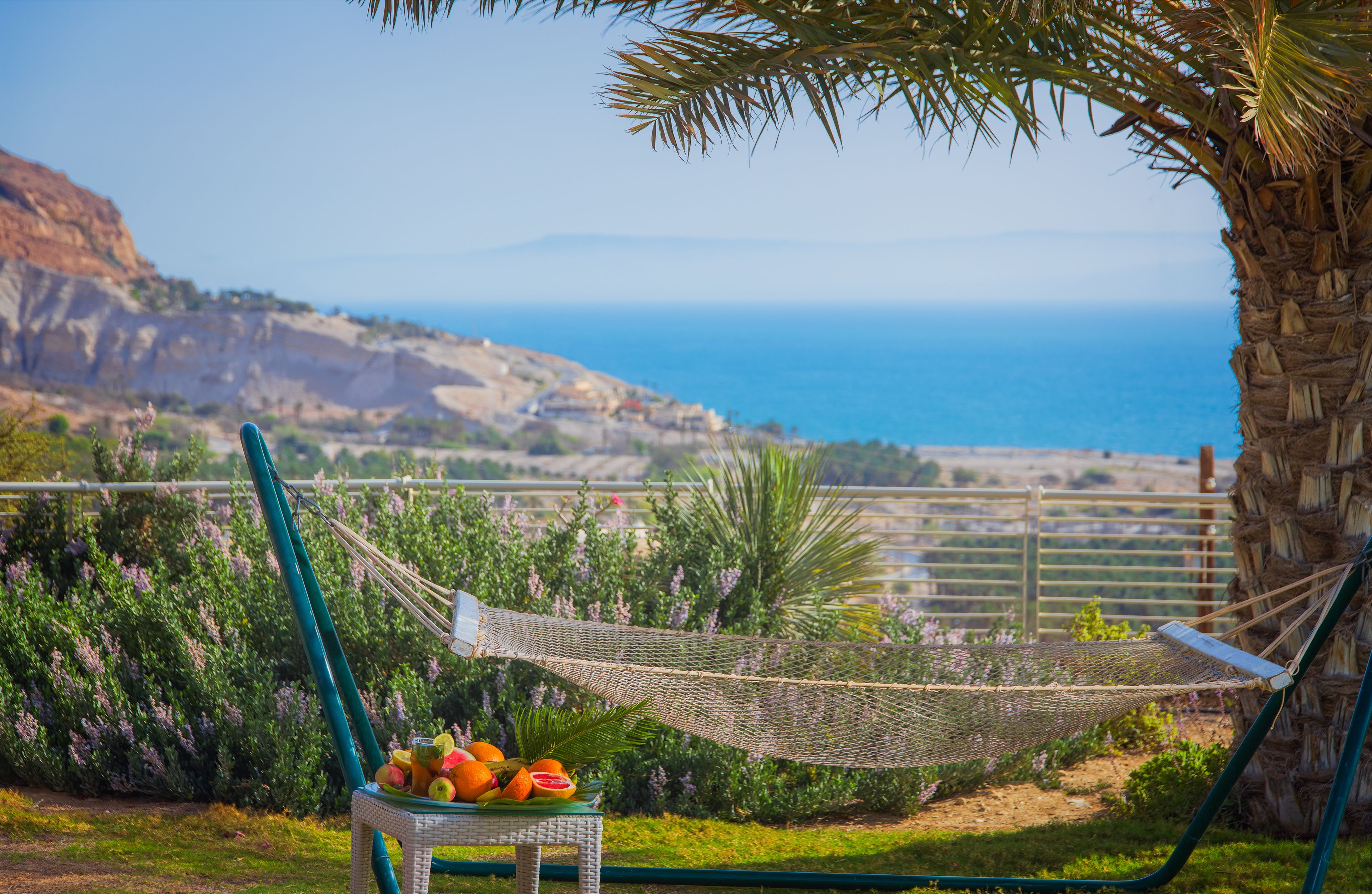 A hammock with a wicker chair and fruit basket, overlooking a coastal landscape with a clear view of the sea and hills.
