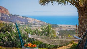 A hammock with a wicker chair and fruit basket, overlooking a coastal landscape with a clear view of the sea and hills.