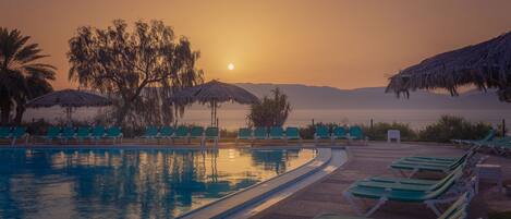 A poolside area with lounge chairs and umbrellas, a clear view of the sunset, and a calm body of water reflecting the sky.