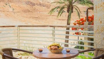 A wooden deck with wicker chairs and a table, overlooking a desert landscape with mountains and a palm tree.