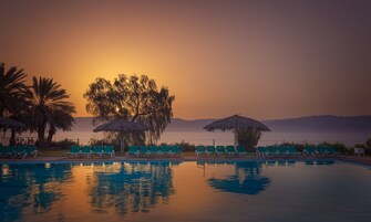 Una piscina con sillas de descanso y sombrillas, rodeada de palmeras y vista al atardecer.