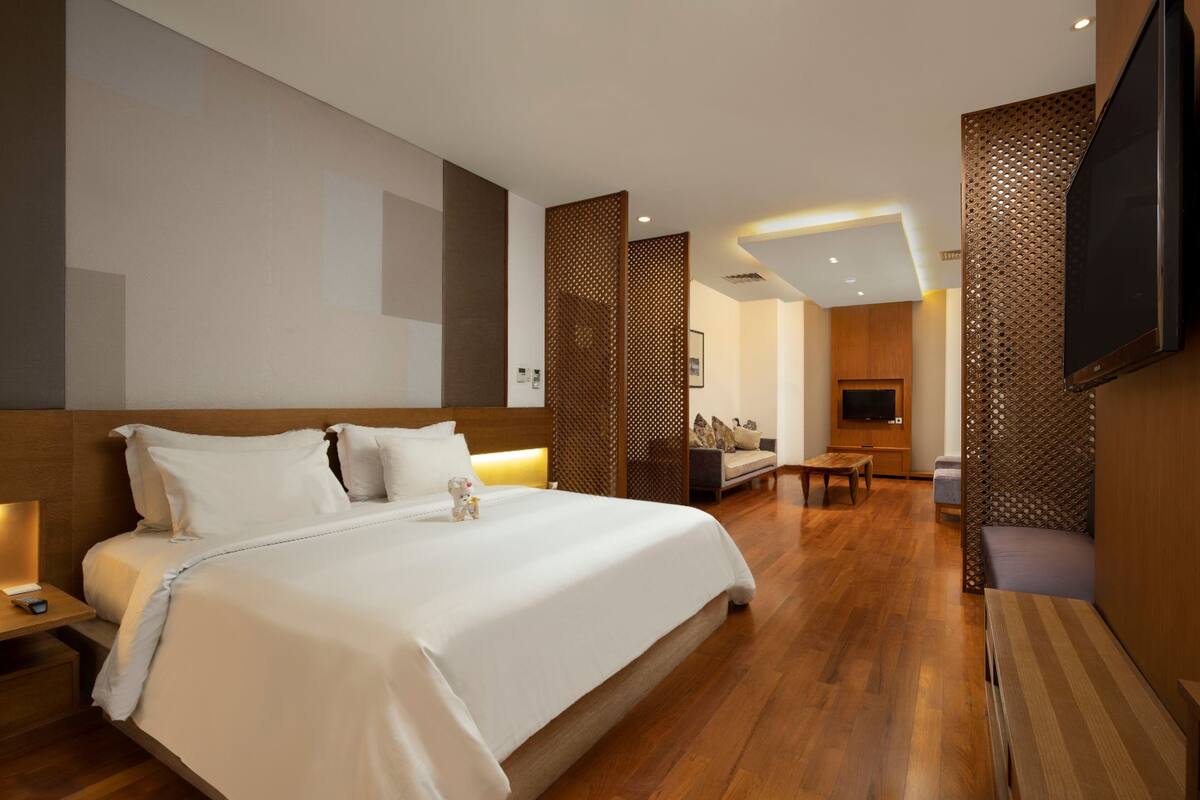 A modern hotel room with a large bed, a TV mounted on the wall, a wooden floor, and a seating area with a sofa and a small table.