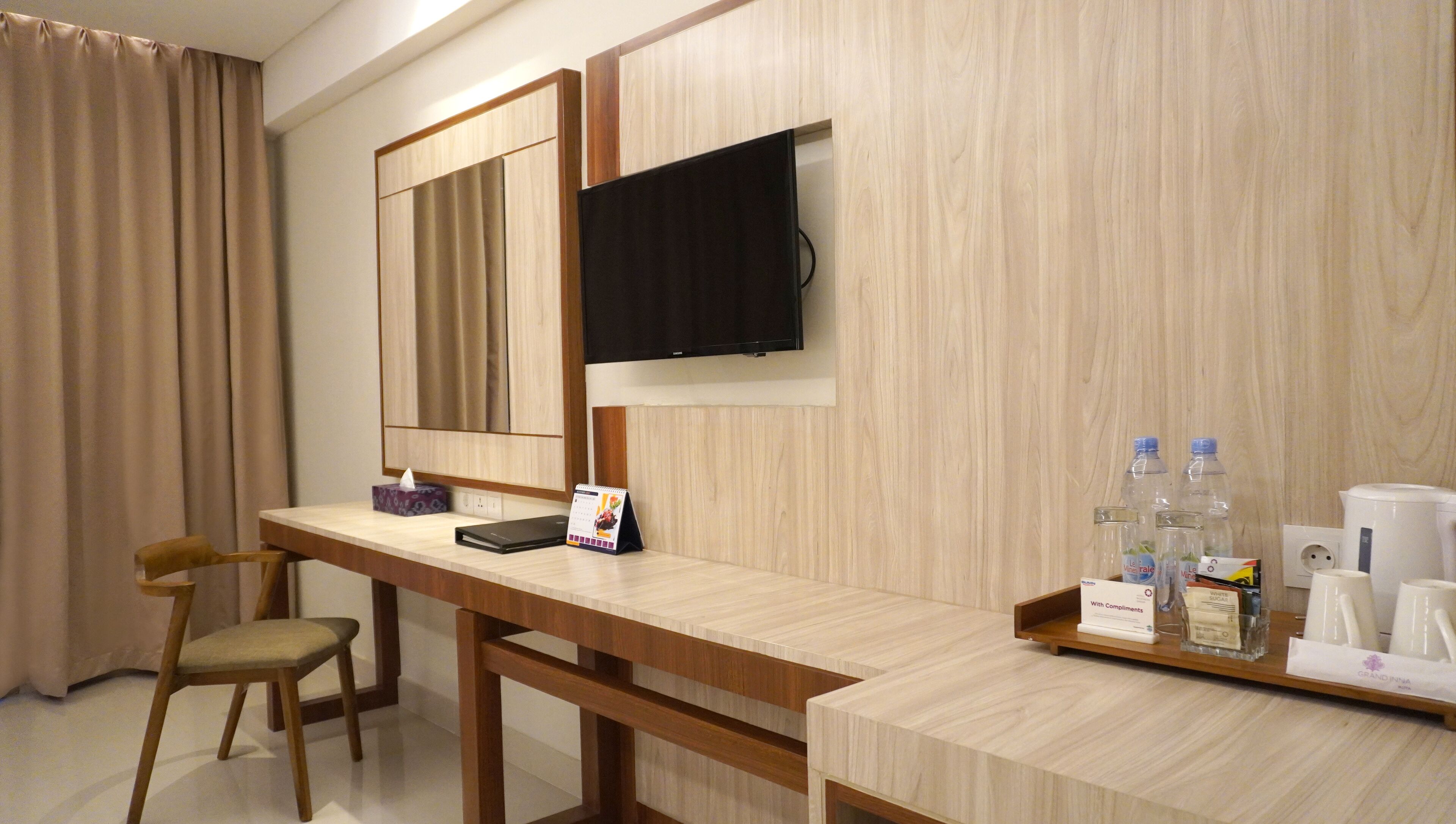 A hotel room with a wooden desk, a chair, a television mounted on the wall, and a tray with toiletries.