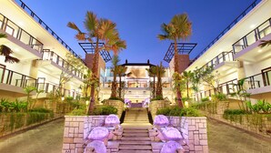 Un hotel con una escalera central flanqueada por palmeras y estatuas decorativas, que conduce a un edificio con varios balcones y un interior bien iluminado.