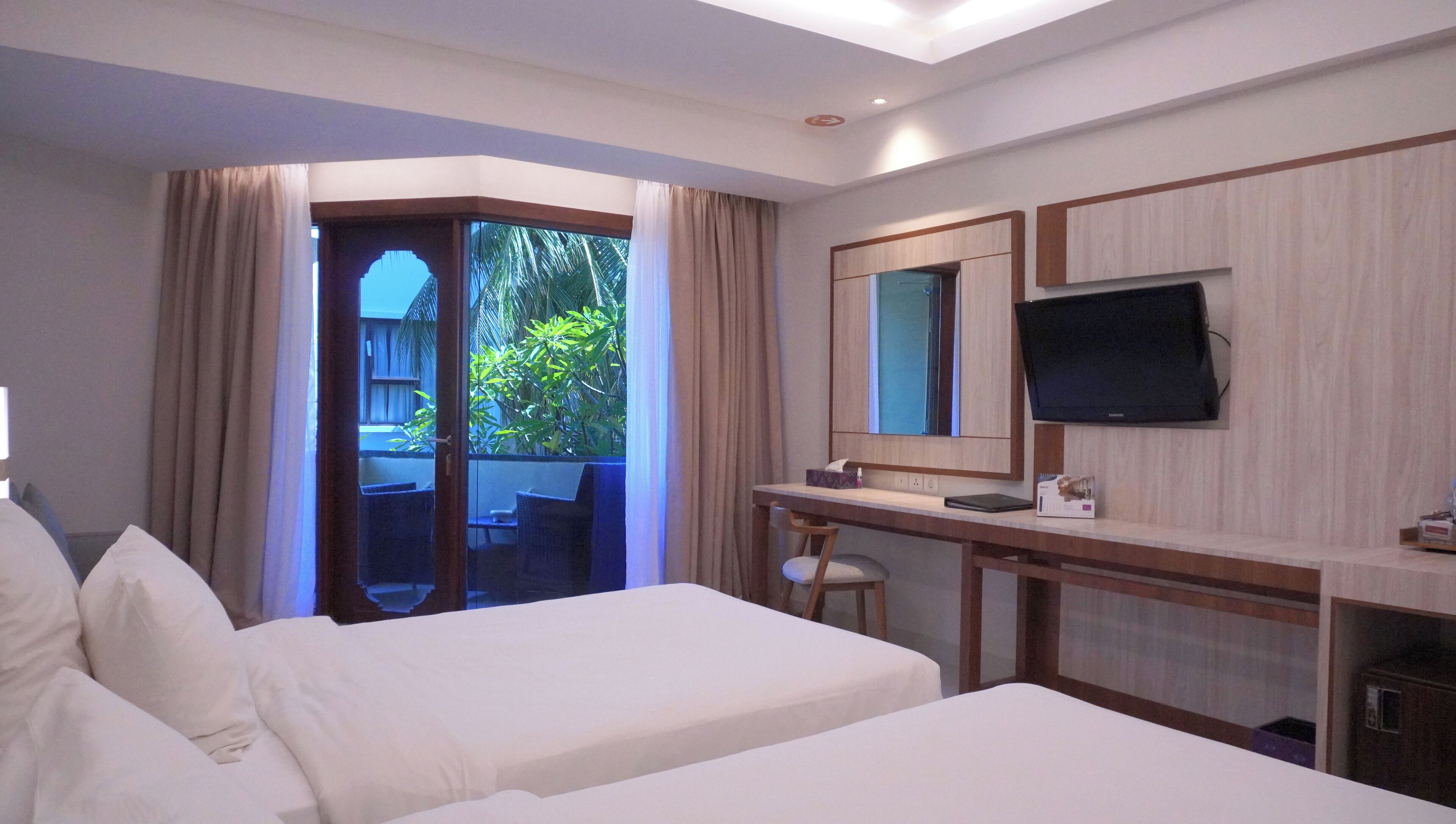 A hotel room with two beds, a desk with a TV, and a balcony with curtains.