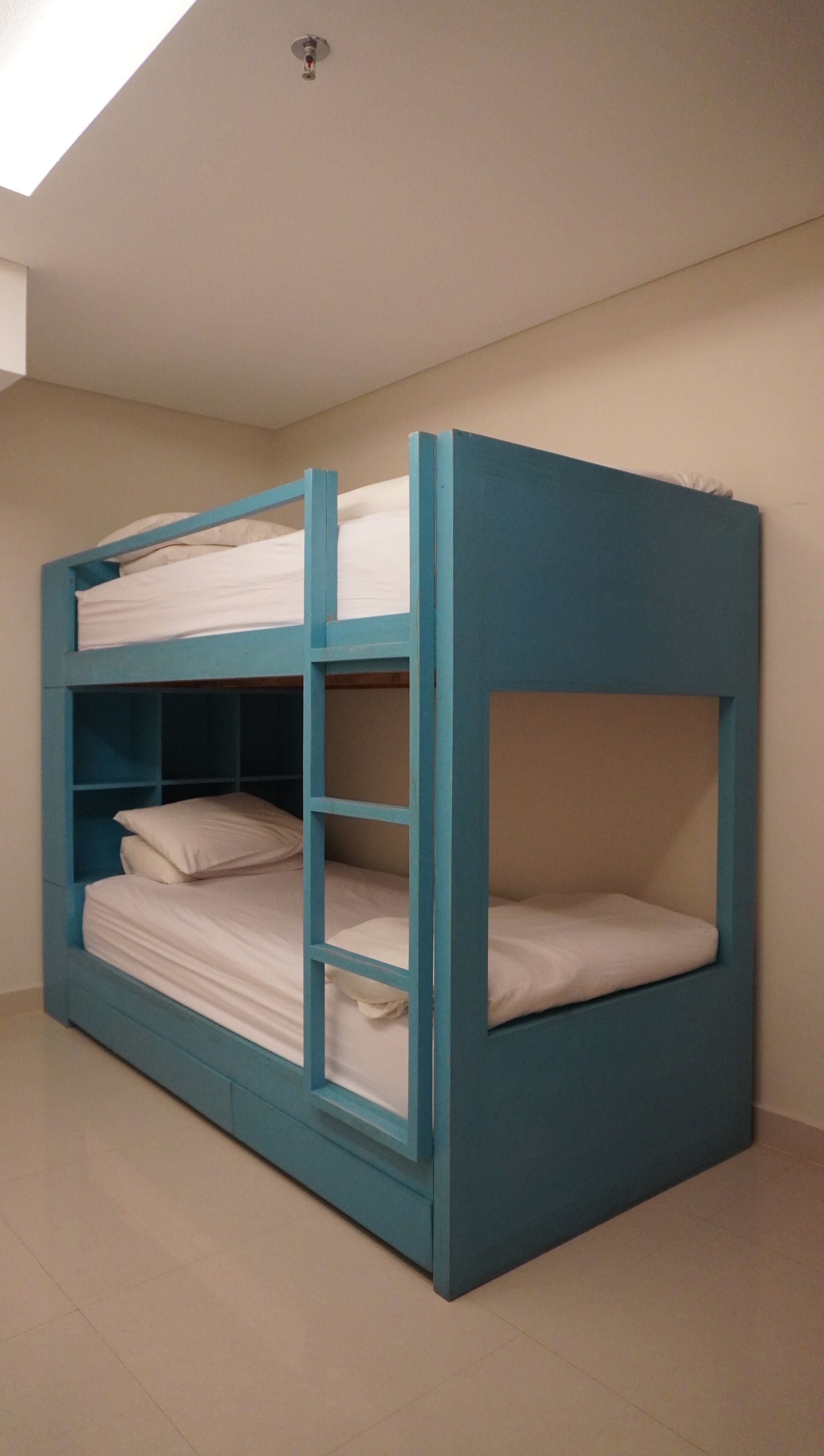 A bunk bed with a blue frame and white bedding in a room with beige walls and a tiled floor.