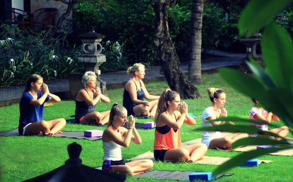 Un grupo de personas sentadas sobre esterillas de yoga al aire libre en un área cubierta de césped, con árboles y un farol de piedra al fondo.