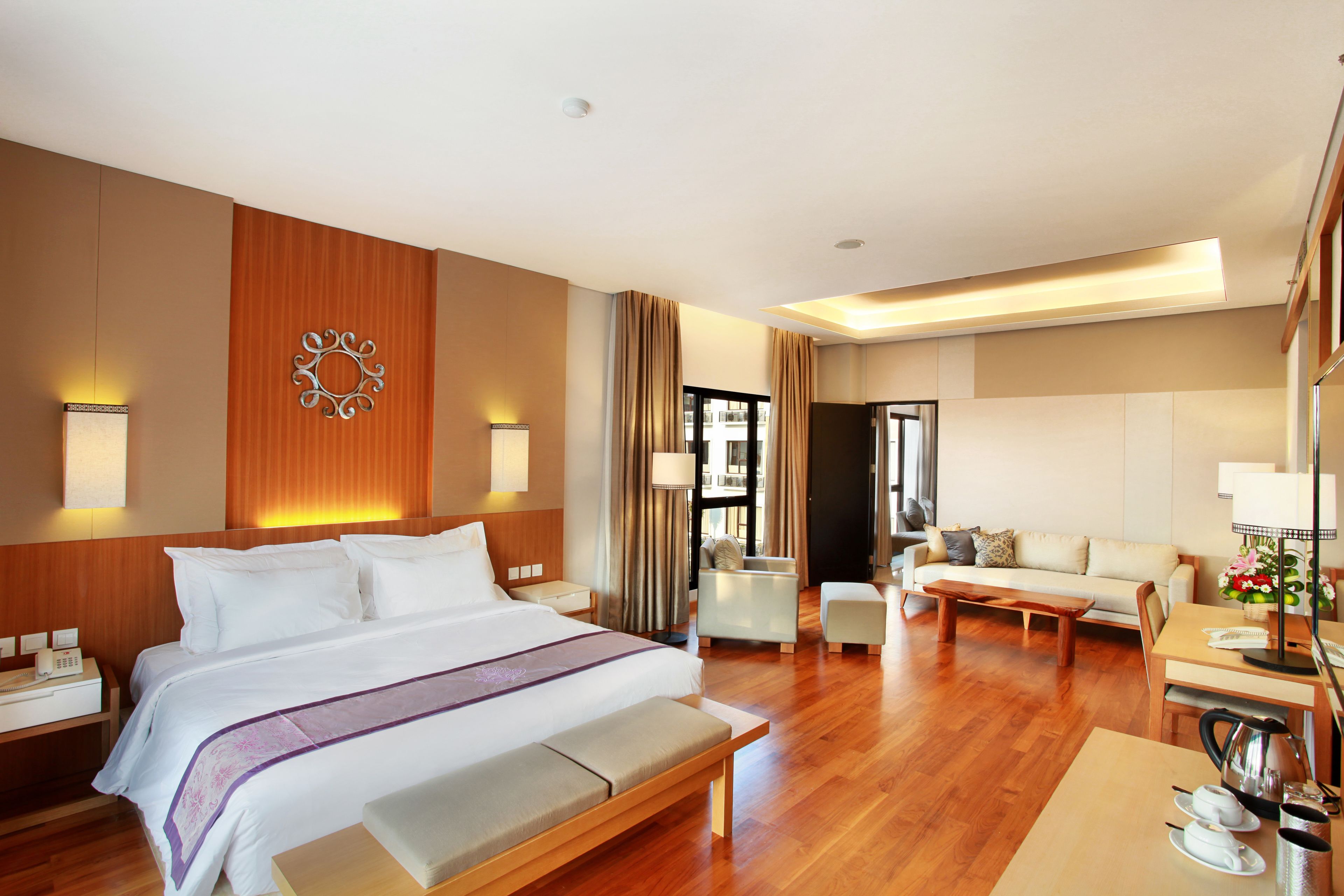 A hotel room with a large bed, a sitting area with a sofa and chairs, a dining table with a kettle, and a view of the outside.