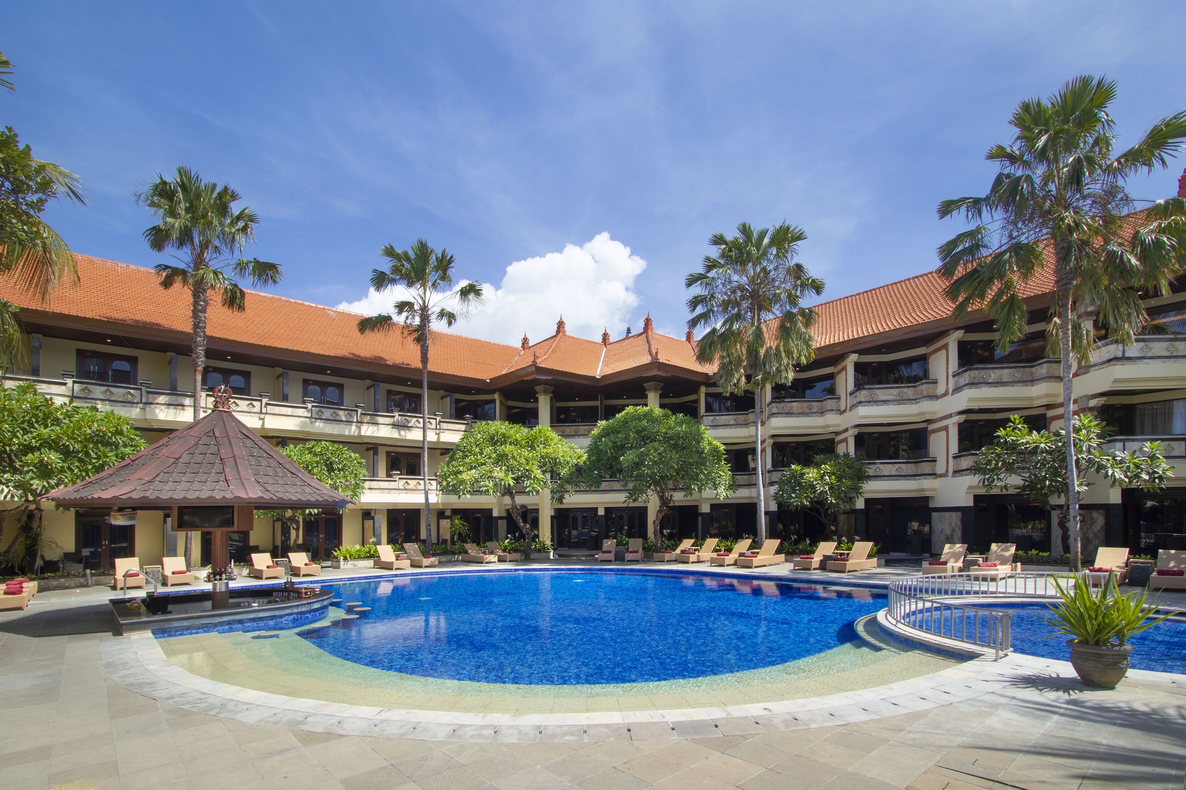 A hotel with a large outdoor swimming pool surrounded by lounge chairs and palm trees.