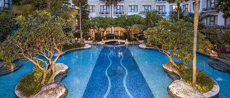 Una piscina de hotel con un elemento central de agua, rodeada de palmeras y otra vegetación tropical.