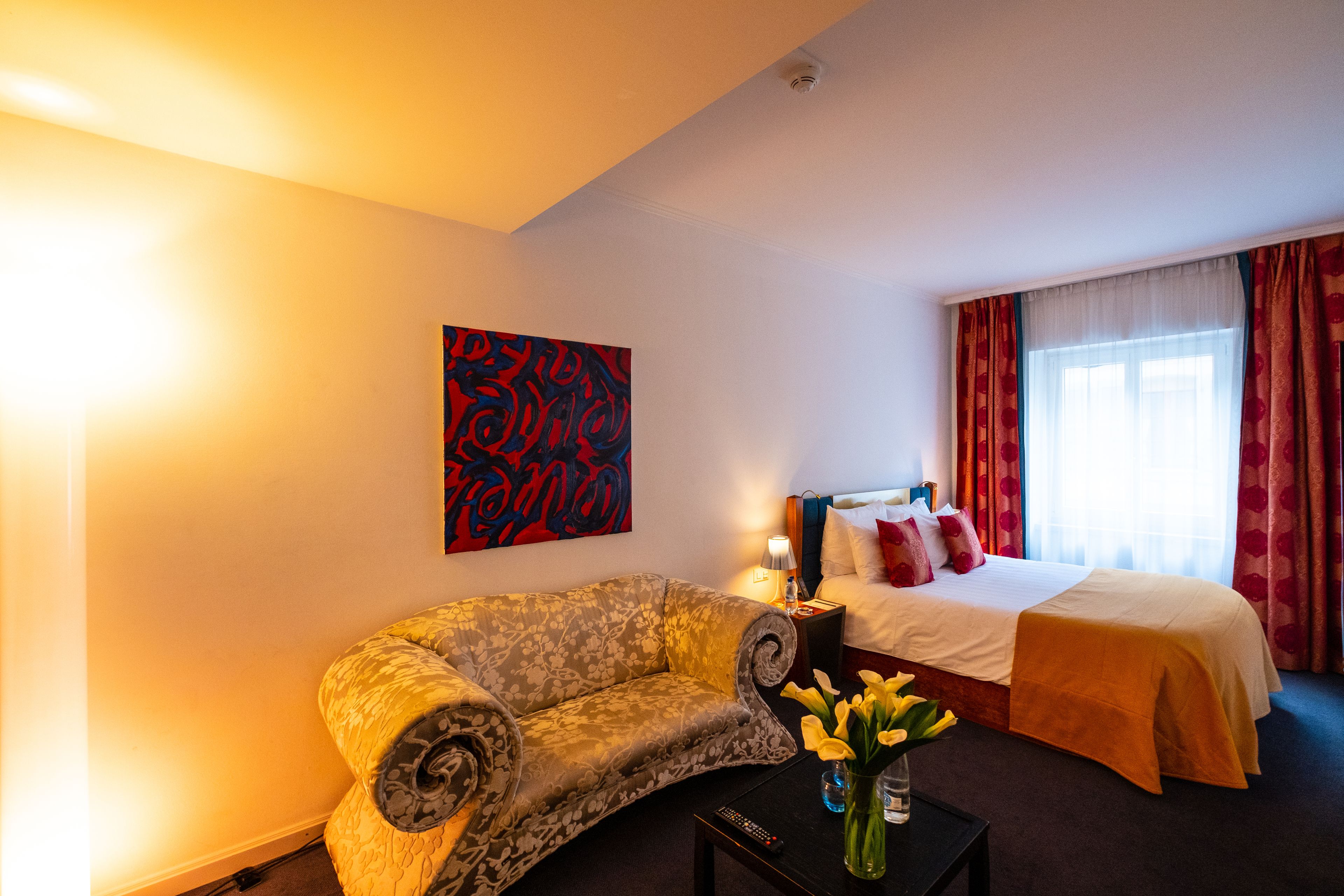 Une chambre d’hôtel avec un canapé, un lit, une table de chevet, une lampe et un vase rempli de fleurs.