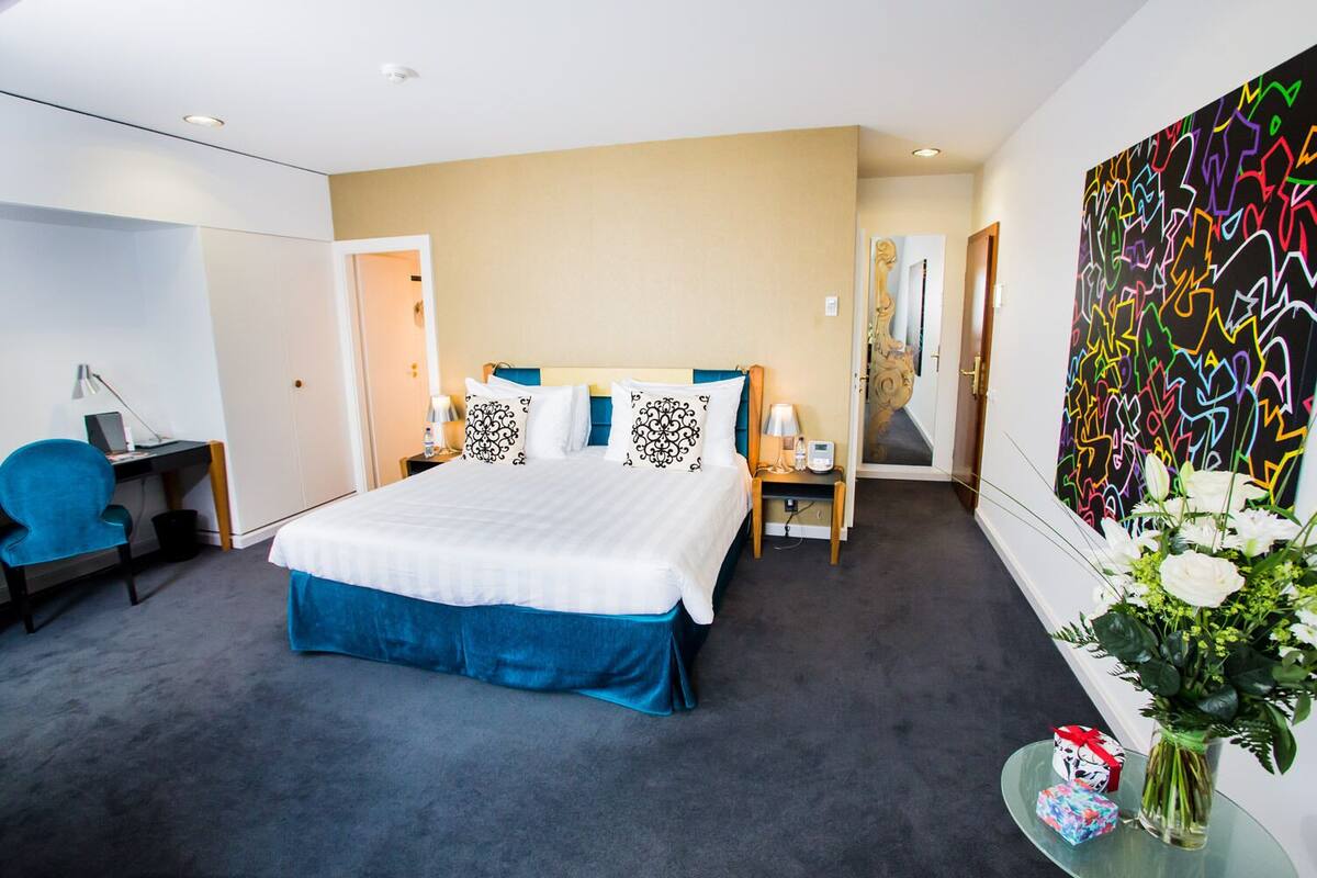 A hotel room with a large bed, a desk with a chair, a small table with a lamp, and a wall with a colorful abstract painting.