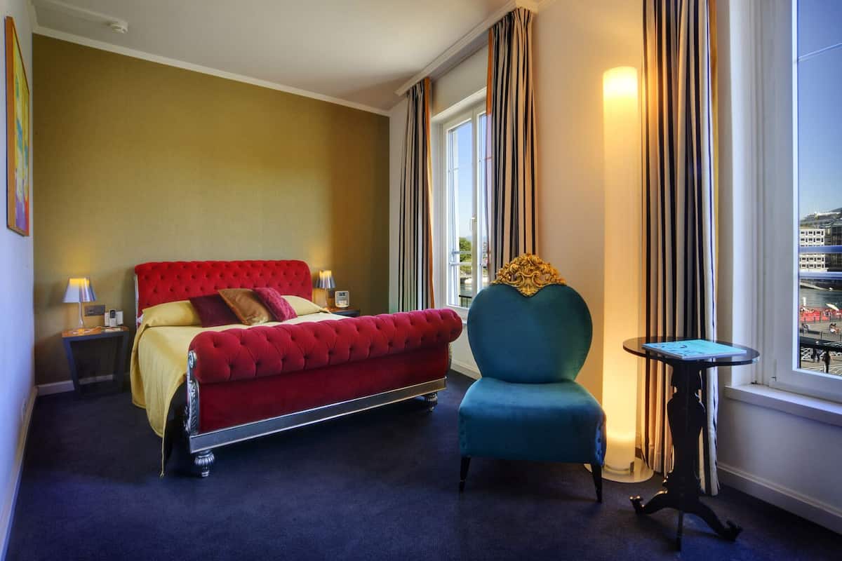 A hotel room with a red upholstered bed, a blue armchair, a small table, and a view of a cityscape through the window.
