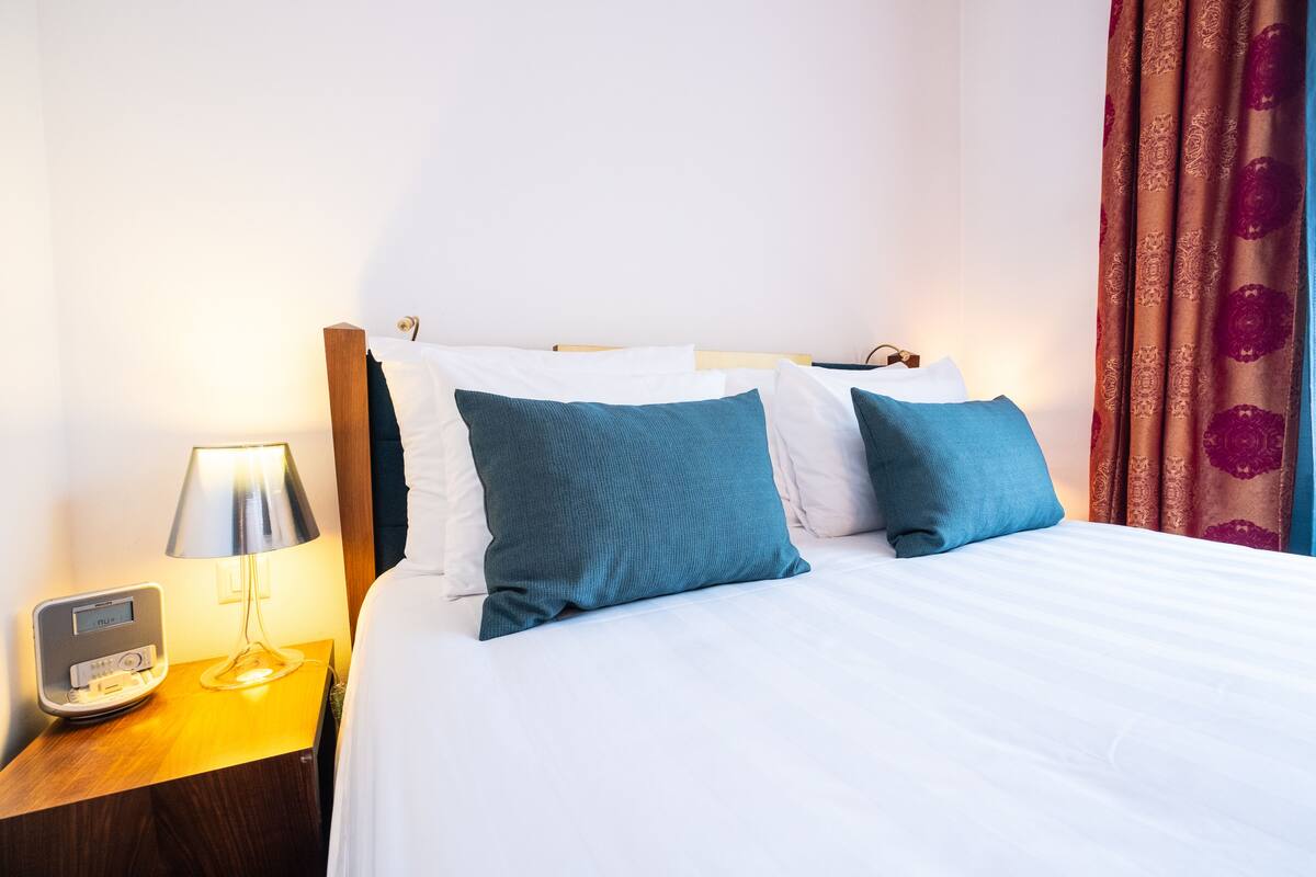 A neatly made bed with white and teal pillows, a bedside table with a lamp, and a digital clock.
