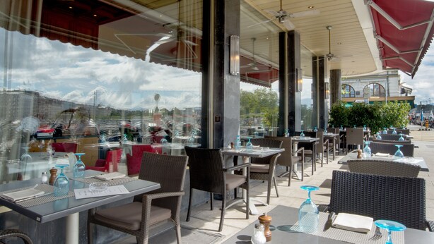 An outdoor cafe with tables and chairs, large windows, and a view of the cityscape.