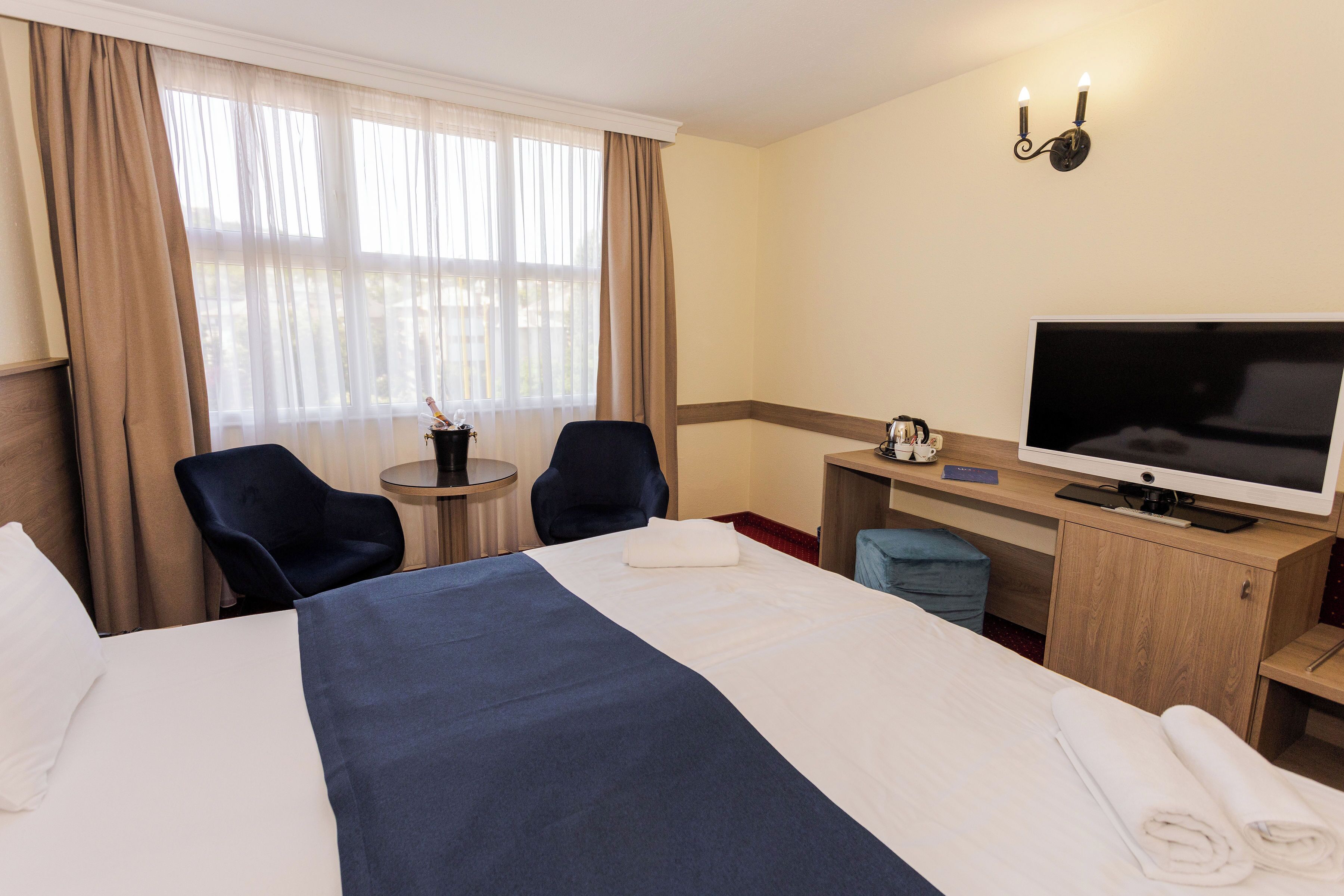 A hotel room with a large bed, a desk with a television, two chairs, a small table with a coffee maker, and a window with curtains.