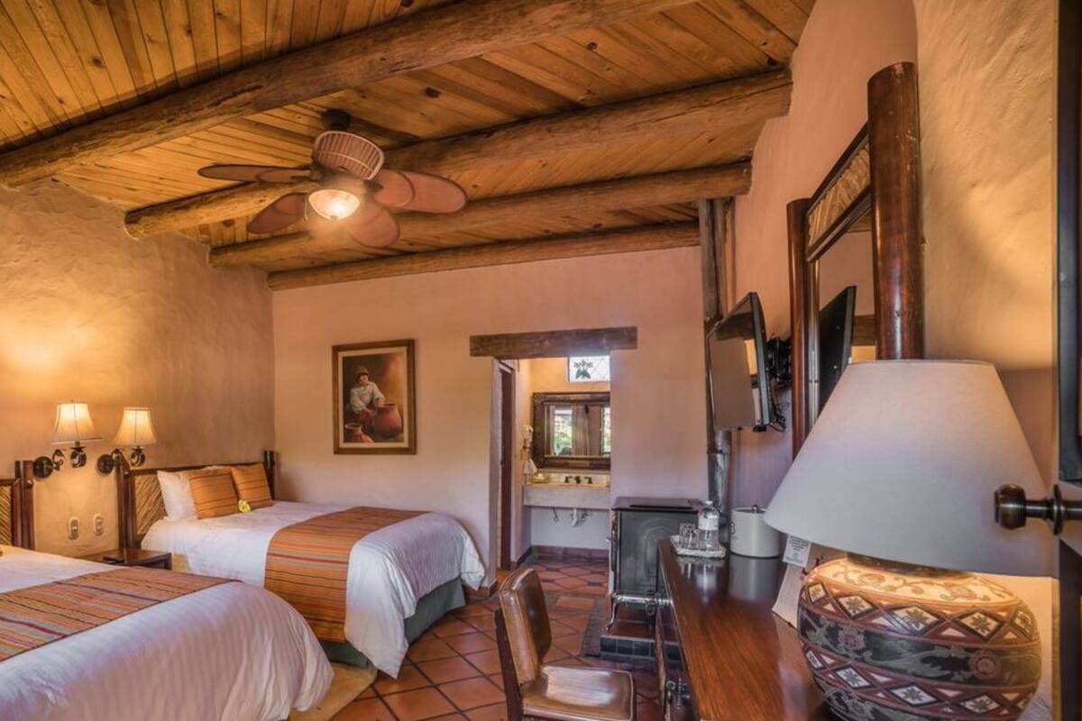 A cozy room with two beds, wooden ceiling beams, a ceiling fan, a fireplace, and a lamp.