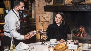 A restaurant setting with a fireplace, a table set for two with wine glasses, a candle, and a menu. - Hotel Mision Cerocahui by Balderrama Hotel Collection (Urique)