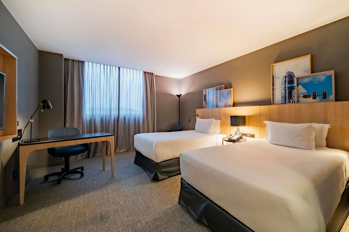 Habitación de hotel con dos camas, un escritorio, una silla, una lámpara y una obra de arte en la pared.