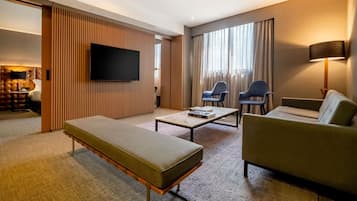 A modern hotel room with a sofa, a small table, a TV, and a large window with curtains.