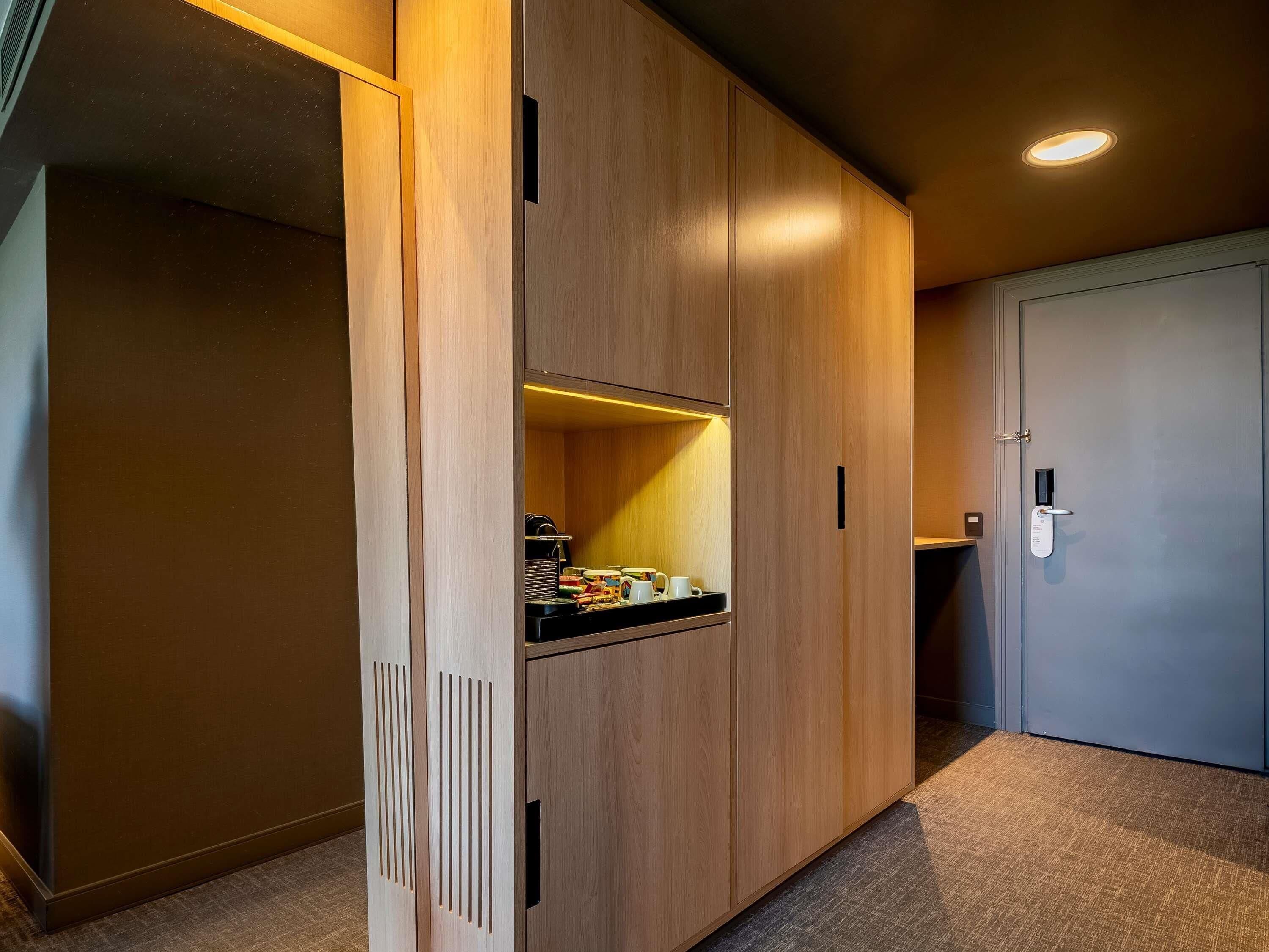A modern hotel room with a wooden wardrobe, a small kitchenette, and a door with a digital lock.