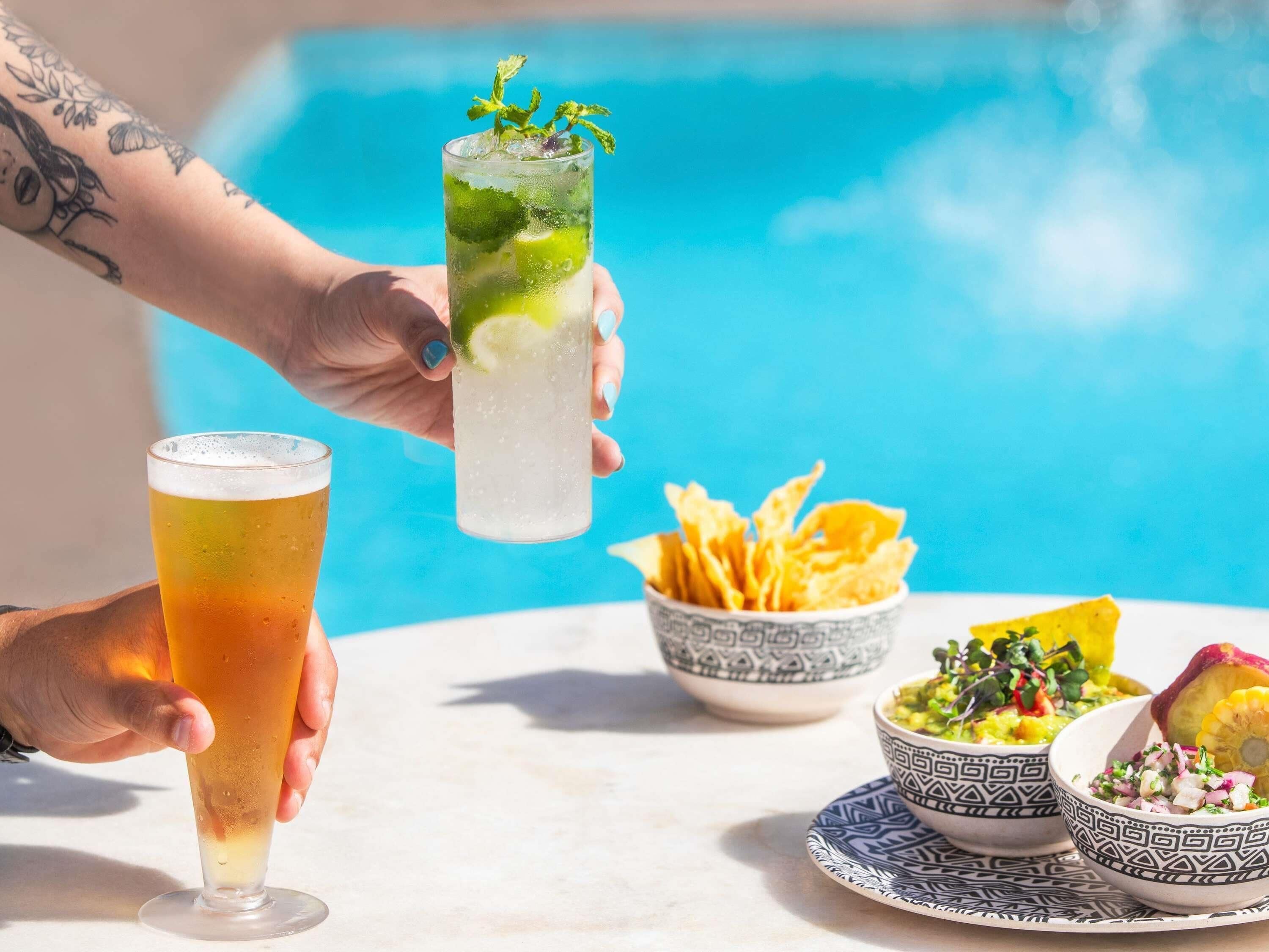 A hand holding a tall glass with a lime and mint garnish, a short glass with a golden liquid, and bowls of food on a table by a pool.