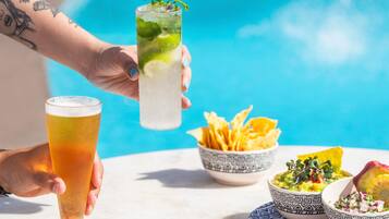 A hand holding a tall glass with a lime and mint garnish, a short glass with a golden liquid, and bowls of food on a table by a pool.
