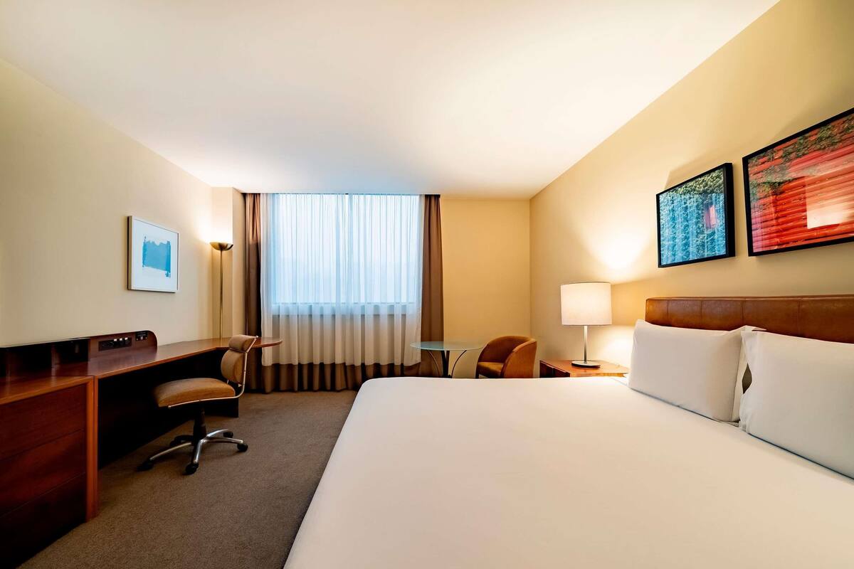 Una habitación de hotel con cama, escritorio, silla, lámpara y una obra de arte en la pared.