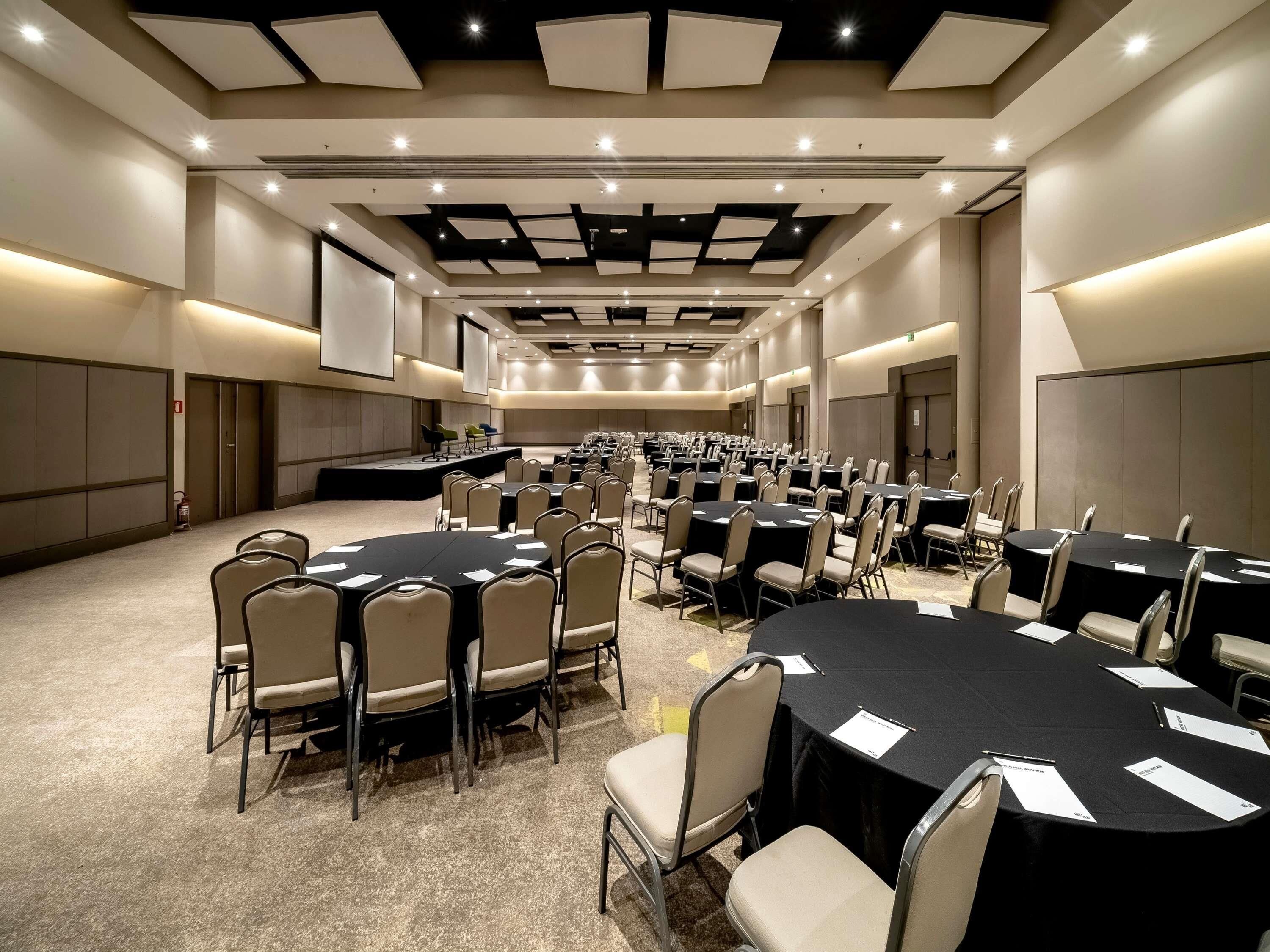 Une grande salle de conférence dotée de nombreuses tables rondes et de chaises, d’une scène et d’un plafond haut avec des luminaires encastrés.