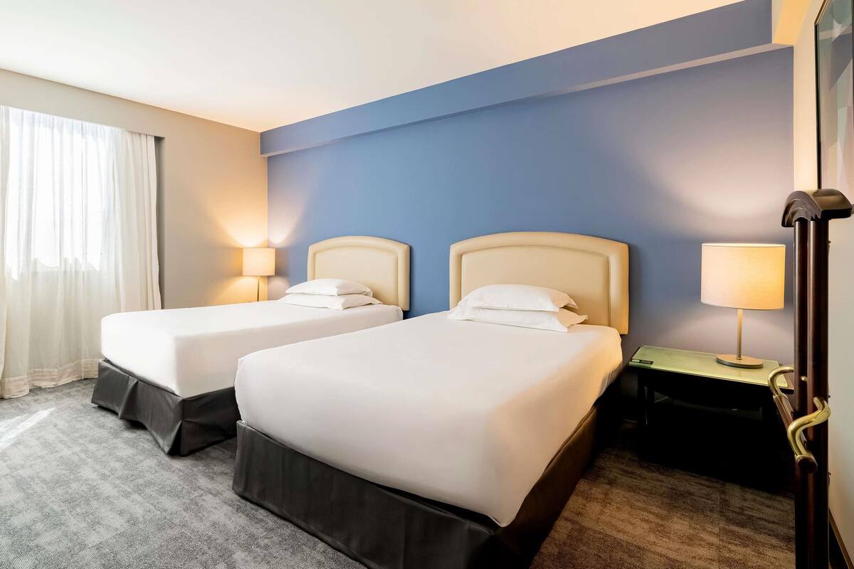 Habitación de hotel con dos camas, una mesita de noche, una lámpara y una ventana con cortinas.